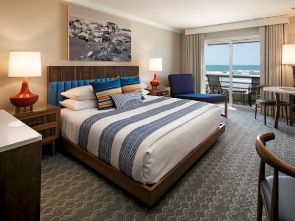 A stylish hotel room with a king bed, blue-striped bedding, nightstands with red lamps, a seating area, and a balcony view of the ocean.