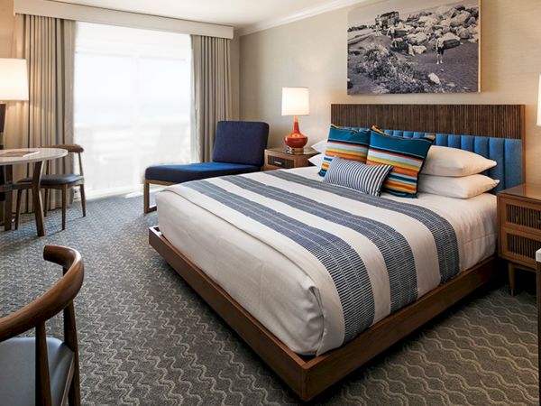 A stylish hotel room with a king bed, blue upholstered headboard, striped bedding, two nightstands with lamps, a seating area, and a desk near a large window.