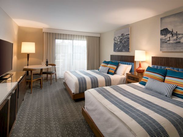 A hotel room with two double beds, striped blue and gray bedding, nightstands with lamps, a desk and chair, a TV stand, and wall art in a softly lit, modern suite.