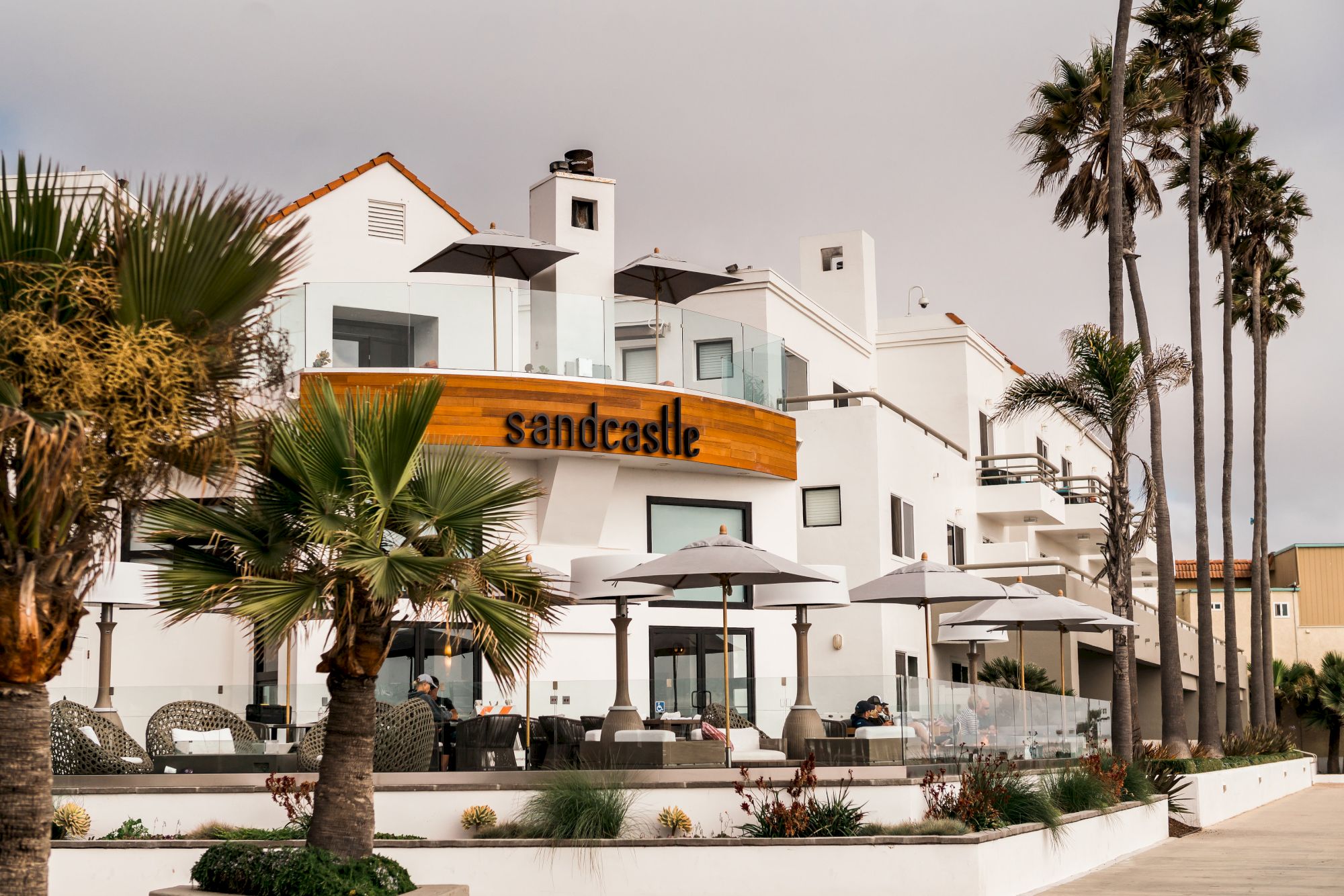 A white, two-story building with an orange sign reading &ldquo;Sonderaste&rdquo; or similar, outdoor seating, umbrellas, palm trees, and a cloudy sky.