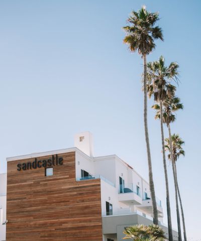 A modern white building with wooden panel accents labeled &ldquo;sandcastle,&rdquo; tall palm trees, and a clear blue sky, sunny coastal vibe.