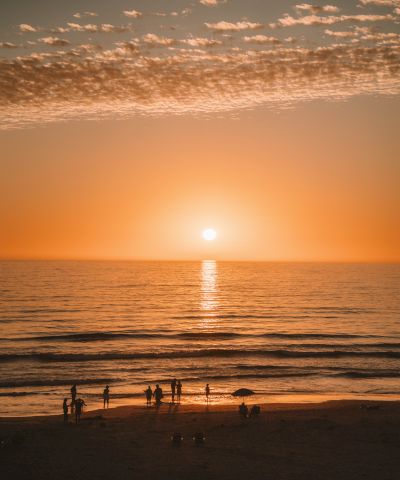 A serene sunset over the ocean with warm orange hues, a calm horizon, and silhouettes of people on the beach enjoying the moment.