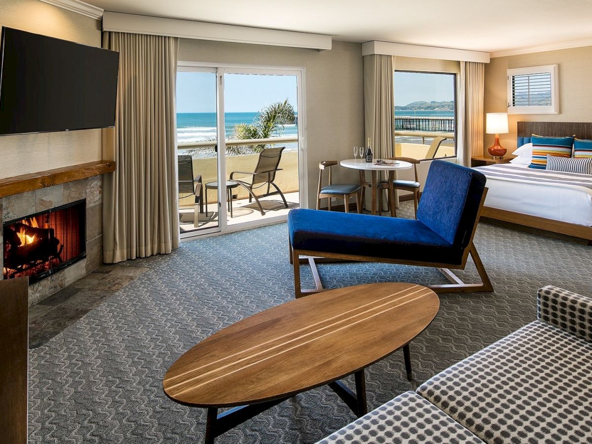 A luxurious hotel suite with a fireplace, seating area, king bed, and balcony overlooking the ocean, bright and modern.