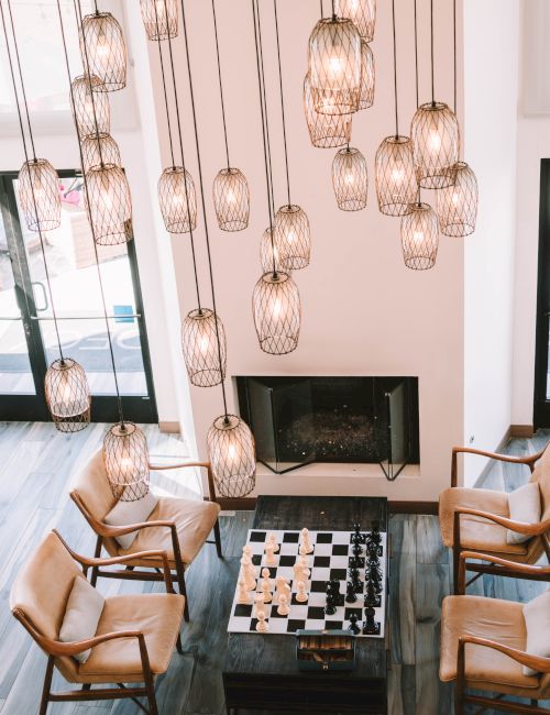 A stylish living room with a high ceiling, pendant cage lights, a chessboard table, and four wooden chairs surrounding it, plus a fireplace in the background.