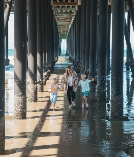 Family walks under a pier, sunlit columns casting long shadows on wet sand, ocean to the side, kids in playful outfits.