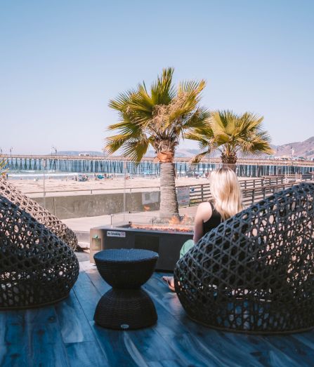 A woman sits on a blue-tiled deck beside two woven basket chairs, facing a sea view with palm trees and a distant pier under clear skies.