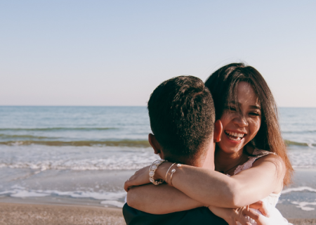 Two people hug on a sunny beach, smiling at the camera, with the ocean and waves in the background.