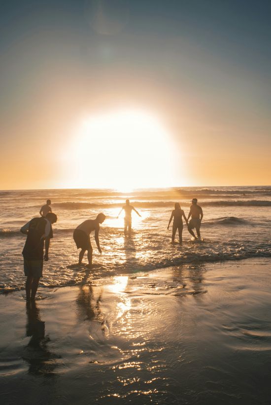 A group of surfers wading and playing in gentle waves at sunset on a beach, silhouettes against a bright, golden horizon.
