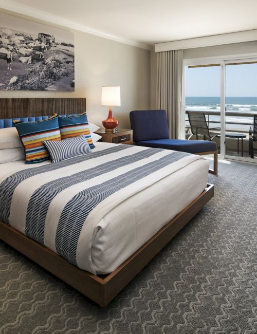 A luxe hotel room with a king bed, striped bedding, nightstands, lamps, a seating area, desk, and a balcony with ocean views.
